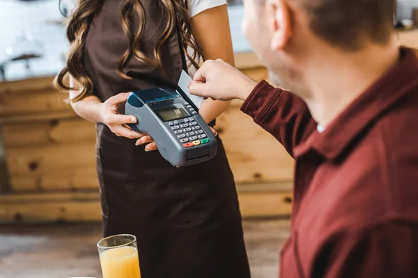 Vista cortada de garçonete segurando terminal wile homem bonito sentado à mesa e pagando com cartão de crédito na casa de café — Fotografia de Stock