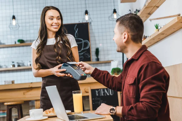 Hermosa camarera sosteniendo terminal wile freelancer sentado en la mesa con el ordenador portátil y pagar con teléfono inteligente en la cafetería - foto de stock