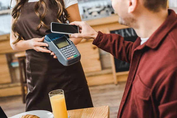 Garçonete vista de barista segurando terminal wile freelancer à mesa pagando com smartphone em cafetaria — Fotografia de Stock