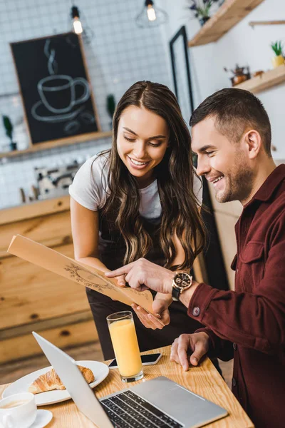 Hermosa camarera y sentado en la mesa hombre guapo mirando al menú en la cafetería — Stock Photo