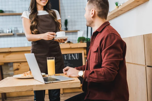 Freelancer sitting at table with laptop wile waitress standing near and holding white cup and saucer in coffee house — Stock Photo