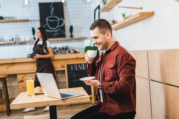 Избирательный фокус красивого фрилансера, сидящего за столом с ноутбуком и пьющего кофе, коварная официантка, стоящая возле барной стойки в кофейне — стоковое фото