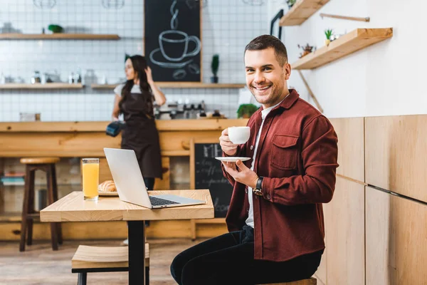 Красивый фрилансер, сидящий за столом с ноутбуком, пьющий кофе и улыбающаяся хитрая официантка, стоящая возле бара в кофейне — стоковое фото