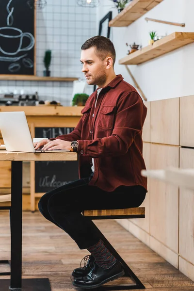 Handsome freelancer in burgundy shirt sitting at table with laptop and typing in coffee house — Stock Photo