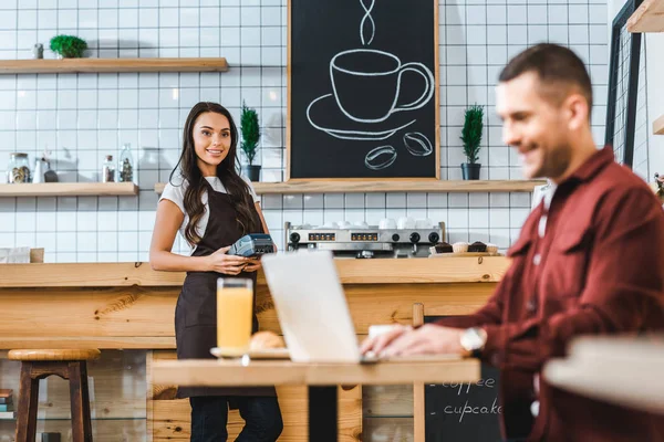 Selective focus of waitress standing near bar counter with terminal wile freelancer in burgundy shirt sitting at table in coffee house — Stock Photo