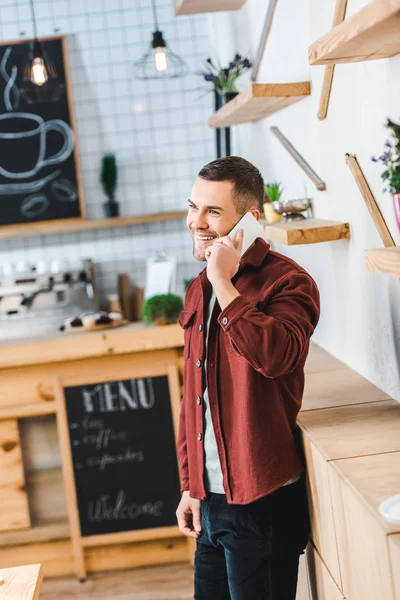 Handsome man in burgundy shirt and jeans standing and talking on smartphone  in coffee house — Stock Photo