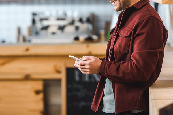 Cropped view of man in burgundy shirt standing with smartphone in coffee house — Stock Photo