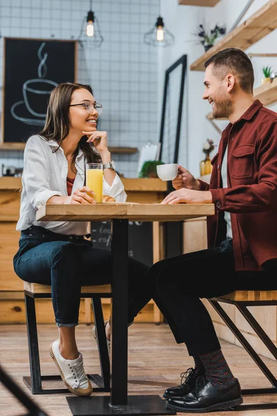 Séduisante femme avec un verre de jus et bel homme avec une tasse de café assis à table dans un café — Photo de stock