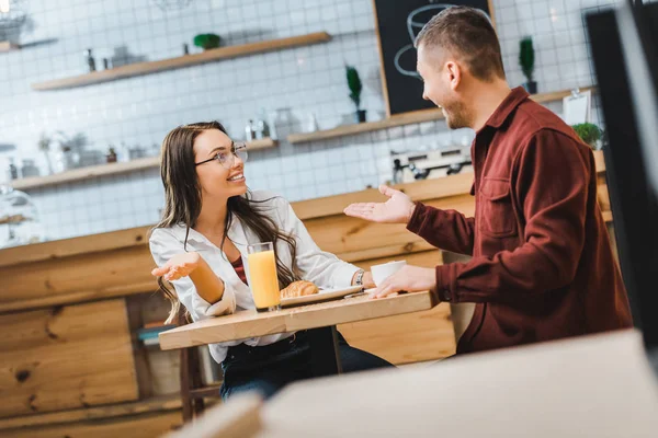 Foco seletivo de mulher morena atraente com vidro de suco e homem bonito com xícara de café sentado e sorrindo à mesa na casa de café — Fotografia de Stock