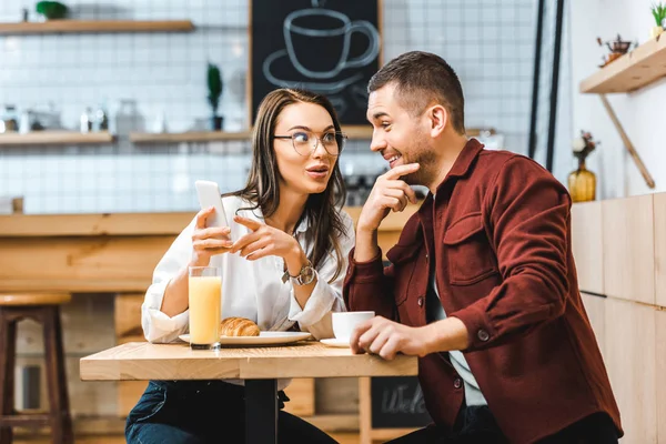 Atractiva mujer morena y hombre guapo sentado en la mesa y mirando al teléfono inteligente en la cafetería - foto de stock