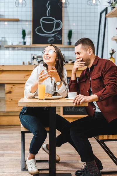 Привлекательная брюнетка и красивый мужчина в бордовой рубашке сидят за столом, смеются и смотрят на смартфон в кофейне — стоковое фото
