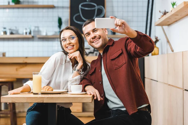 Привлекательная брюнетка и красивый мужчина в бордовой рубашке сидят за столом и берут селфи на смартфон в кофейне — стоковое фото