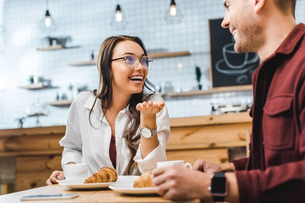 Atractiva mujer morena y hombre guapo en camisa de color burdeos sentado a la mesa con cruasanes, sonriendo y hablando en la cafetería - foto de stock