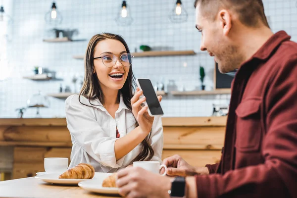 Привлекательная брюнетка, сидящая за столом и показывающая смартфон с чистым экраном красивому мужчине в бордовой рубашке в кофейне — стоковое фото