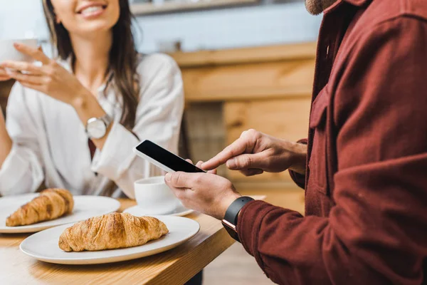 Vista recortada de hombre sosteniendo teléfono inteligente con pantalla en blanco mujer astuta sentado a la mesa y sonriendo en la cafetería - foto de stock