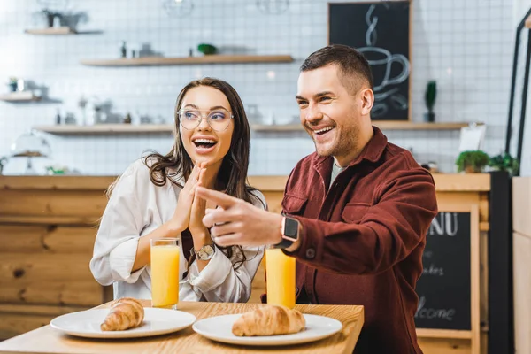 Mulher morena atraente e homem bonito em camisa borgonha sentado e sorrindo à mesa com croissants e copos de suco na casa de café — Fotografia de Stock