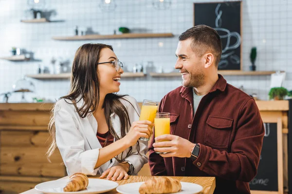 Mulher morena atraente e homem bonito em camisa borgonha sentado à mesa com croissants, sorrindo e clinking óculos com suco na casa de café — Fotografia de Stock