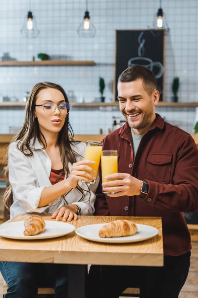 Beautiful brunette woman and handsome man sitting at table with croissants and clinking glasses with juice in coffee house — Stock Photo
