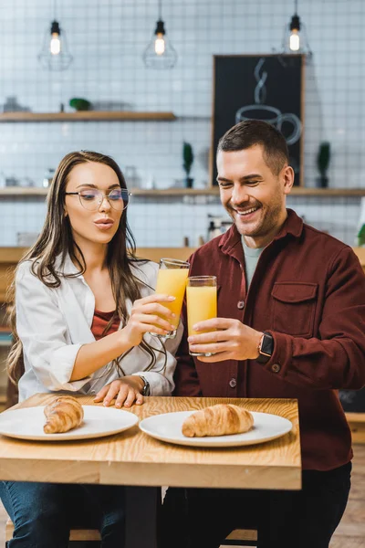 Hermosa mujer morena y hombre guapo sentado a la mesa con croissants y tintineo vasos con jugo en la cafetería - foto de stock