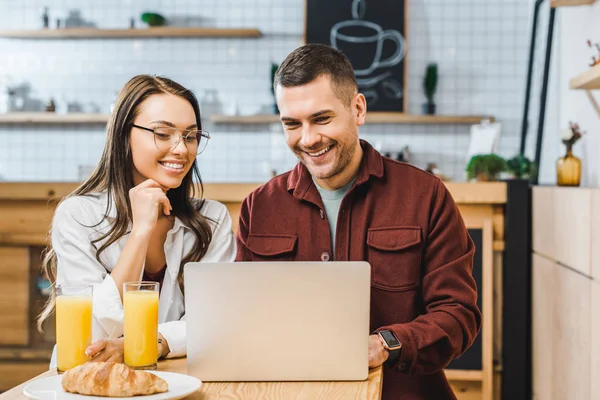 Attractive brunette woman and handsome man sitting at table and looking to laptop in coffee house — Stock Photo