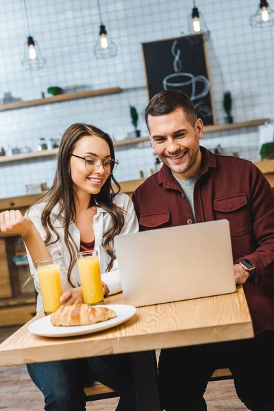 Bella donna bruna e bell'uomo seduto a tavola, sorridente e in cerca di laptop in caffetteria — Foto stock