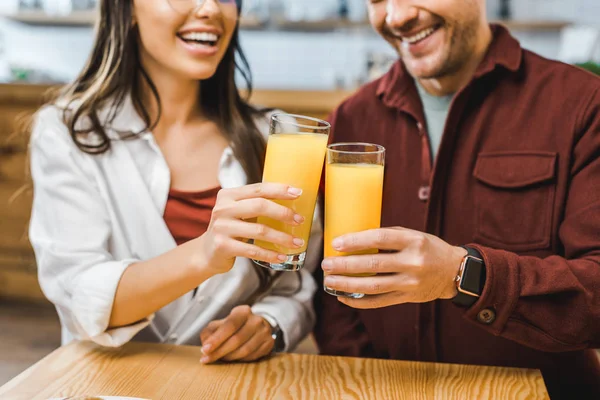 Vista recortada de la mujer y el hombre sentado a la mesa y tintineo vasos con jugo en la cafetería - foto de stock