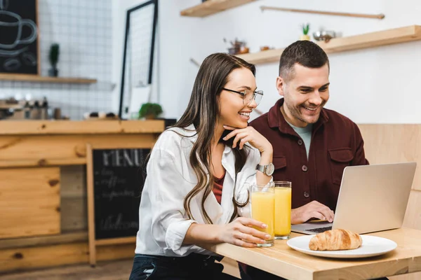 Beautiful brunette woman and handsome man sitting at table with juice in glasses and croissants and looking to laptop in coffee house — Stock Photo