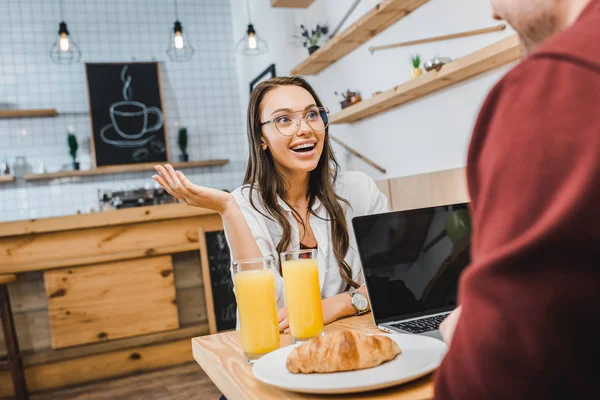 Selective focus of attractive woman sitting at table with croissant and juice in glasses and talking to man with laptop in coffee house — Stock Photo