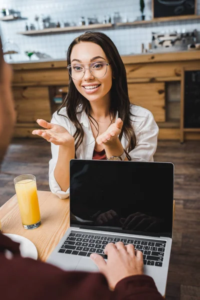Selective focus of attractive woman sitting at table with juice in glass and talking to man with laptop in coffee house — Stock Photo