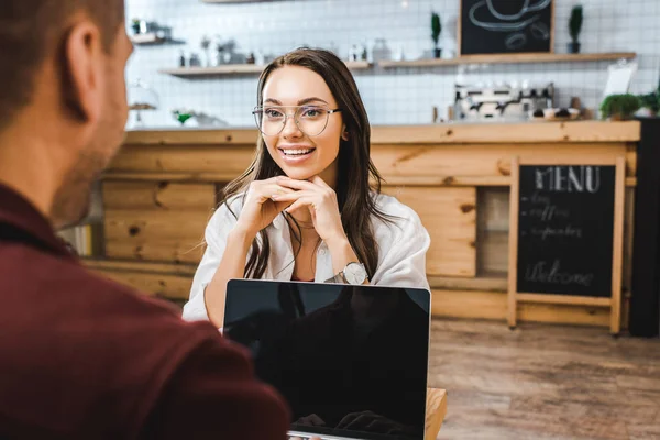 Selective focus of attractive woman sitting at table and talking to man with laptop in coffee house — Stock Photo