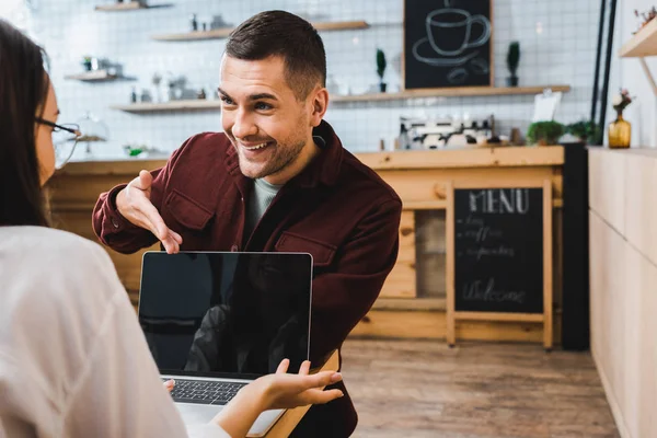 Schöner Mann im weinroten Hemd sitzt am Tisch und zeigt auf Laptop mit Hand gegenüber Frau im Kaffeehaus — Stockfoto