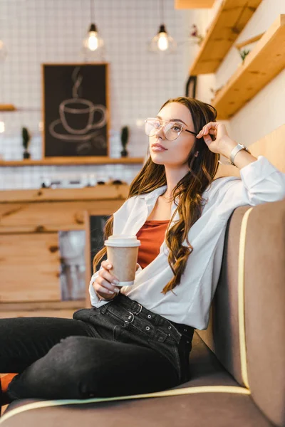 Attractive woman in white shirt and black jeans sitting on couch and holding paper cup in coffee house — Stock Photo