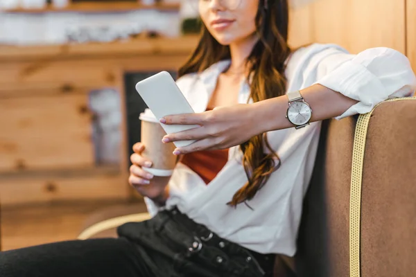 Vista recortada de la mujer en camisa blanca y vaqueros negros sentados en el sofá, sosteniendo la taza de papel y el teléfono inteligente en la cafetería - foto de stock