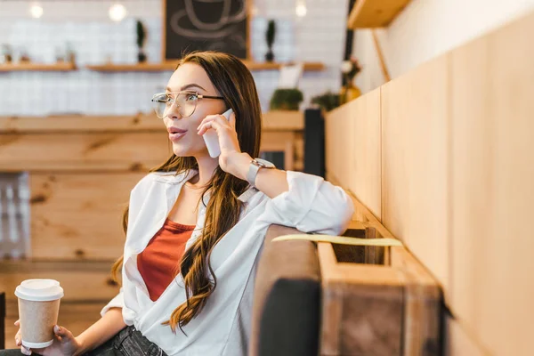 Attraente donna in camicia bianca seduta sul divano, tenendo in mano la tazza di carta e parlando sullo smartphone in caffetteria — Foto stock