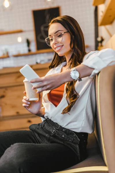 Mulher atraente em camisa branca sentado no sofá, segurando copo de papel e olhando para smartphone na casa de café — Fotografia de Stock