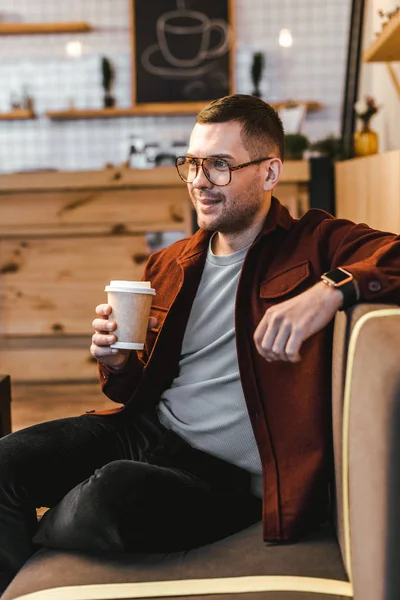 Man in burgundy shirt and black jeans sitting on couch and holding paper cup in coffee house — Stock Photo