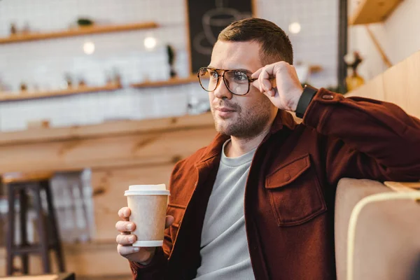 Schöner Mann in weinrotem Hemd und schwarzer Jeans sitzt auf der Couch und hält Pappbecher im Kaffeehaus — Stockfoto