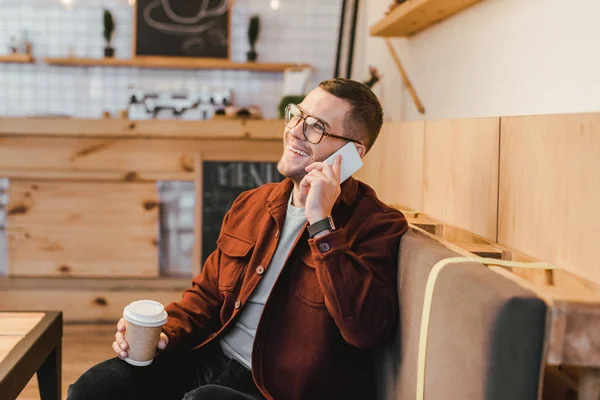 Man in burgundy shirt, black jeans sitting on couch, holding paper cup and talking on smartphone in coffee house — Stock Photo