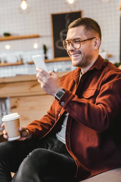 Man in burgundy shirt sitting on couch, holding paper cup, looking to smartphone and laughing in coffee house — Stock Photo