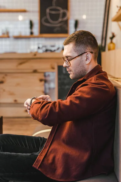 Handsome man in burgundy shirt and black jeans sitting on couch and touching watch with fingein coffee house — Stock Photo