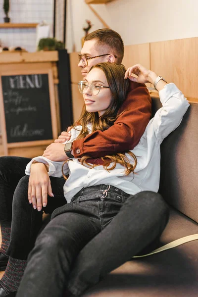 Atractiva mujer y hombre en gafas sentados en el sofá y abrazos en la cafetería - foto de stock