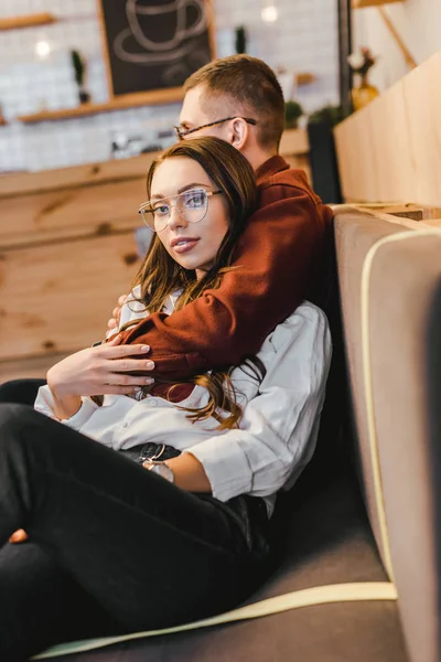 Attraente donna in camicia bianca e uomo in occhiali seduto sul divano e abbraccio in caffetteria — Foto stock