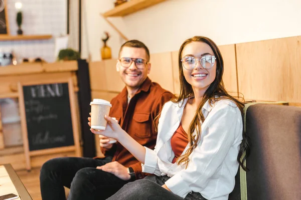 Attraente donna in possesso di tazza di carta e seduto sul divano con l'uomo in camicia bordeaux in caffetteria — Foto stock