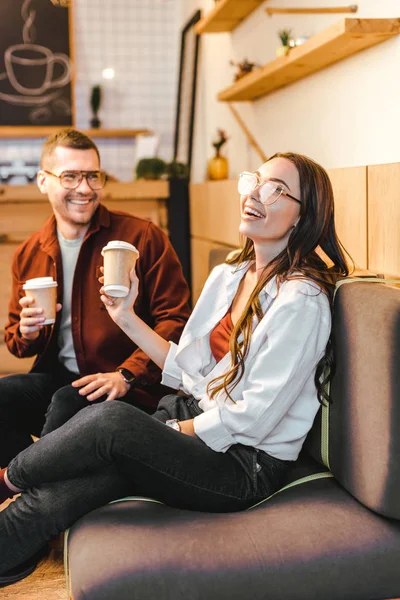 Donna attraente e bell'uomo seduto sul divano, con in mano tazze di carta e ridere in caffetteria — Foto stock