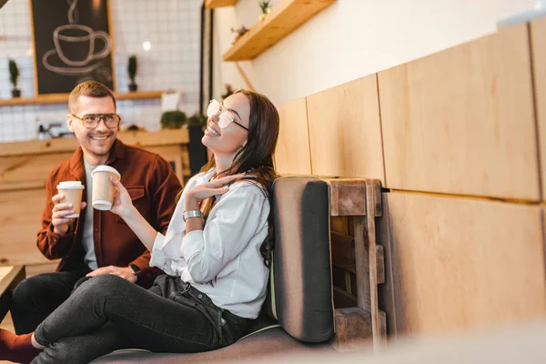 Donna attraente e bell'uomo seduto sul divano, con in mano tazze di carta e ridere in caffetteria — Foto stock