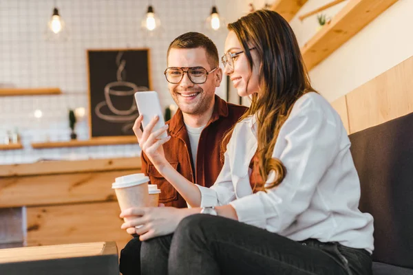 Attractive woman and handsome man sitting on couch, holding paper cups and looking to smartphone in coffee house — Stock Photo