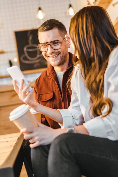 Morena mujer y hombre guapo sentado en el sofá, sosteniendo vasos de papel y mirando a teléfono inteligente en la cafetería - foto de stock