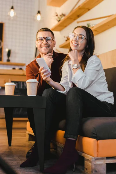 Attractive woman holding smartphone and sitting on couch with handsome man at table with paper cups in coffee house — Stock Photo