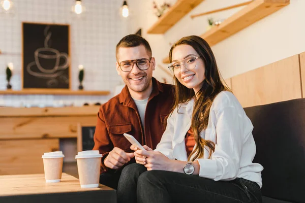 Atractiva mujer sonriendo, sosteniendo el teléfono inteligente y sentado en el sofá con un hombre guapo en la mesa con tazas de papel en la cafetería - foto de stock