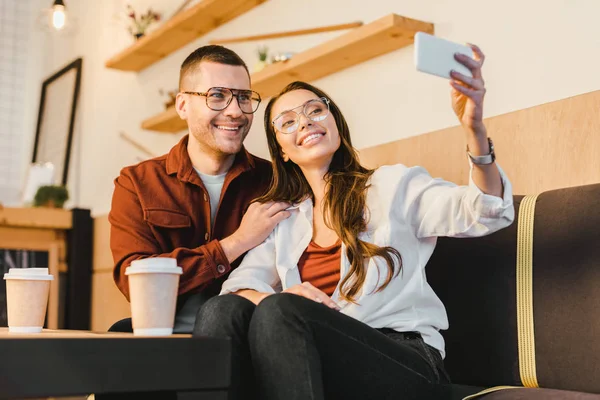 Attraktive Frau und gutaussehender Mann sitzt auf der Couch, lächelt und macht Selfie am Tisch mit Pappbechern im Kaffeehaus — Stockfoto